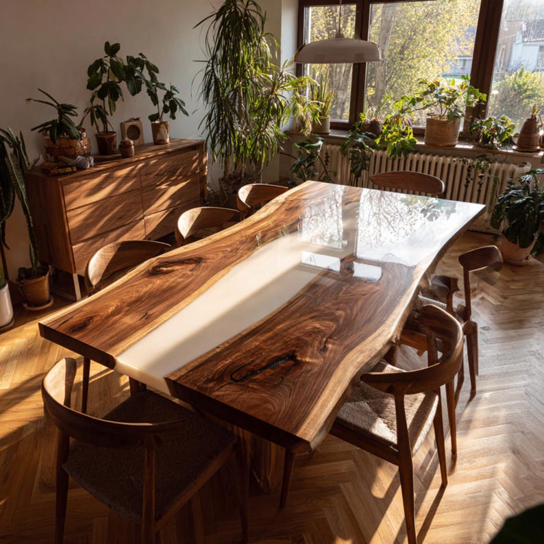 Op de afbeelding: Een grote, handgemaakte houten eettafel met een lichtgekleurd epoxyhars middenstuk. De tafel heeft een live edge design en is omringd door houten stoelen. De kamer is gevuld met natuurlijk licht en planten, wat een warme en uitnodigende sfeer cre&euml;ert.
