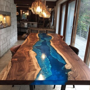 May include: A large, rectangular dining table with a unique design. The table features a dark brown wooden frame with a vibrant blue resin river running through the center. The table is illuminated by hanging lights.