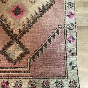 Op de afbeelding: Een geweven tapijt met een oudroze centraal veld en een groot, donkerbruin geometrisch ruitmotief. De rand is lichtbeige met herhalende kleine bloem- en ruitpatronen in tinten van felroze, blauwgroen en donkerbruin.
