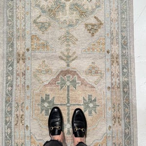 May include: A rectangular area rug featuring a faded, intricate pattern in muted shades of sage green, dusty rose, and soft gold on a cream-colored background. The design includes geometric motifs and a decorative border along the edges.