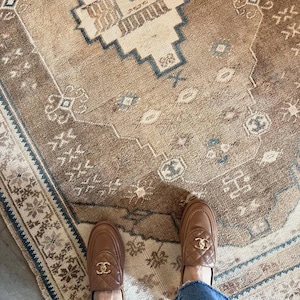 May include: A pair of tan leather loafers featuring a quilted vamp, a rounded toe, and a gold-toned interlocking CC logo emblem on the front. The shoes are worn with blue denim jeans, positioned on a brown and beige patterned area rug.