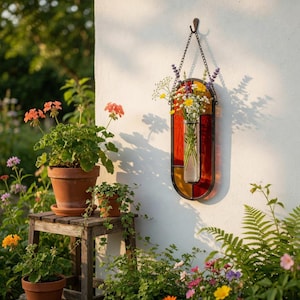 Könnte beinhalten: Ein Wandvase aus Buntglas mit einer Metallkette und einem Haken, gefüllt mit frischen Blumen. Die Vase hat rote, gelbe und orangefarbene Glaspaneele. Sie hängt an einer weißen Wand neben Topfpflanzen und einem hölzernen Pflanzenständer.