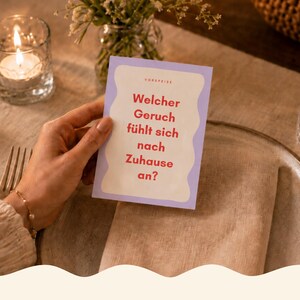 Könnte beinhalten: Ein Dinner-Hosting-Bundle mit einer Karte, die fragt: "Welcher Geruch fühlt sich nach Zuhause an?" Das Bild zeigt eine brennende Kerze, ein Glas Wasser und ein Gedeck. Der Text "DINNER HOSTING BUNDLE" ist rot dargestellt.