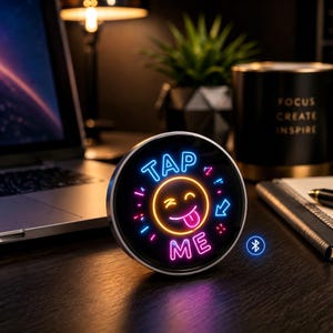 Puede incluir: Un dispositivo de pantalla digital circular con un gráfico estilo neón de una cara sonriente guiñando un ojo con la lengua fuera, rodeado por el texto "TAP ME" en azul y rosa, con un pequeño icono de Bluetooth azul en la esquina inferior derecha.