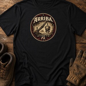 May include: A black short-sleeve t-shirt featuring a circular graphic on the chest. The design depicts a construction crane lifting a bucket with the text ARRIBA. LET'S RAISE IT UP and the initials AR centered within the circular frame.