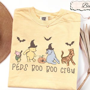 Könnte beinhalten: Ein buttergelbes T-Shirt mit einer Grafik von Ferkel, Winnie Puuh, I-Aah und Tigger, die Hexenhüte tragen, begleitet von einem Kürbis und Fledermäusen. Der Text PEDS BOO BOO CREW ist in Braun unter den Figuren aufgedruckt.
