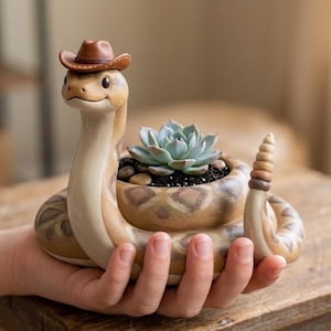 May include: A tan and brown rattlesnake-shaped planter featuring a coiled body, a raised head wearing a small brown cowboy hat, and a central circular opening containing a green succulent plant surrounded by small dark pebbles.