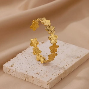 May include: A gold-colored bracelet featuring a floral design. The open cuff bracelet is adorned with multiple flower-shaped embellishments. The jewelry is displayed on a textured, off-white surface against a neutral background.