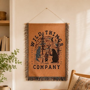 Könnte beinhalten: Ein rechteckiger, gewebter Wandteppich in warmem Orange mit Fransenrand und einer hölzernen Aufhängestange. Das Design zeigt eine Lagerfeuerszene mit einem blauen Wolf und einem schwarzen Bären, umgeben von Bäumen und dem Text Wild Things and Company.