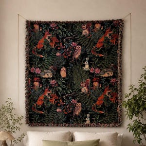 May include: A rectangular woven wall tapestry featuring a dark black background with a repeating pattern of orange foxes, brown owls, squirrels, rabbits, hedgehogs, mushrooms, and green ferns. The tapestry has a fringed edge and is hung by a string.