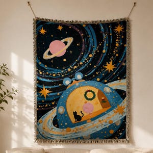 May include: A rectangular woven throw blanket featuring a dark blue space scene with yellow stars, a pink ringed planet, and a large blue planet with a transparent dome. Inside the dome, a black cat stands near a pink creature and a door.
