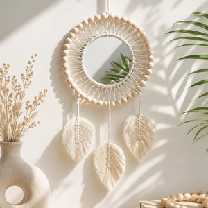 May include: A circular wall mirror framed by light-colored wooden beads and white macrame cord. Three macrame leaf-shaped tassels hang from the bottom of the circular frame. The mirror is suspended by a single white cord attached to the top center.