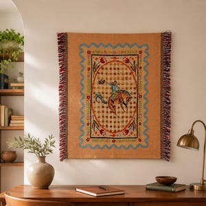 May include: A rectangular woven wall tapestry in a warm tan color featuring a central oval frame with a checkered pattern. Inside the oval, a figure rides a bucking horse. The border is decorated with floral motifs and a blue wavy line.