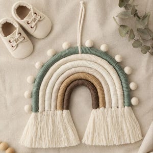 May include: A wall hanging shaped like a rainbow, constructed from wrapped yarn in shades of sage green, cream, tan, and dark brown. The arch features small white felt pom-poms along the top edge and long, cream-coloured fringe at both ends.