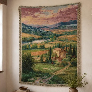 Könnte beinhalten: Ein rechteckiger, gewebter Wandteppich mit einer Landschaftsdarstellung einer Landvilla, umgeben von grünen Feldern, hohen Zypressen und fernen Bergen unter einem rosa-orangen Sonnenuntergangshimmel, versehen mit einem gefransten Rand.
