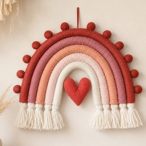 May include: A wall hanging featuring a rainbow made of wrapped yarn in shades of red, pink, and white. The rainbow is decorated with small red pom-poms along the outer arch, a central red fabric heart, and white yarn tassels at both ends.