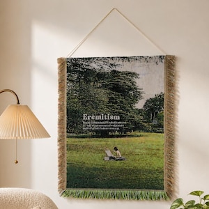May include: A rectangular woven wall hanging featuring a landscape scene of a green field and trees with the word Eremitism printed in white text. The tapestry has a fringed border in shades of brown and green and hangs from a thin white cord.