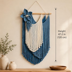 Könnte beinhalten: Ein Makramee-Wandbehang mit blauen und cremefarbenen Baumwollkordeln, die an einem Holzstab hängen. Eine große blaue Blumenapplikation ist auf der linken Seite befestigt. Das Stück misst 50 cm in der Breite und 120 cm in der Höhe.