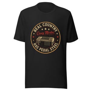 Può includere: T-shirt nera con un design circolare. Il design include il testo "Real Country", "Casey Martin" e "Has Pedal Steel", insieme a un'illustrazione di una chitarra pedal steel in stile vintage.