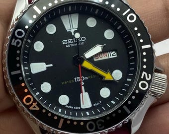 May include: A Seiko automatic wristwatch with a black dial, luminous circular hour markers, and a yellow arrow-shaped second hand. The watch features a black rotating bezel, a day-date window, and a vibrant magenta silicone strap.
