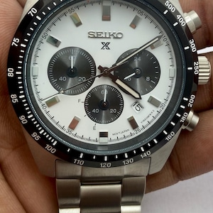 May include: A silver-toned stainless steel Seiko chronograph watch with a white dial, three dark grey sub-dials, and a black tachymeter bezel. The watch features luminous hour markers, a date window at the four o'clock position, and a metal link band.