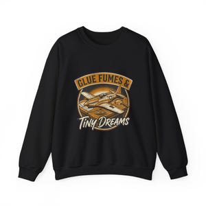 May include: A black crewneck sweatshirt featuring a graphic illustration of a model airplane on a workbench with tools. The text reads GLUE FUMES & TINY DREAMS in a circular orange and brown frame. The design includes a magnifying glass and glue.