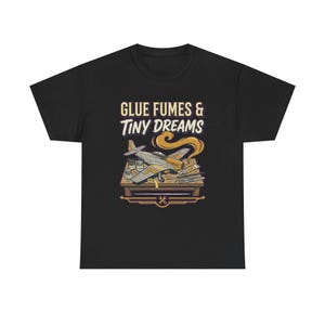 May include: A black t-shirt featuring a graphic illustration of a model airplane on a workbench with paint jars, glue, and tools. The text above the illustration reads GLUE FUMES & TINY DREAMS in a stylized font, with a small wrench icon below.