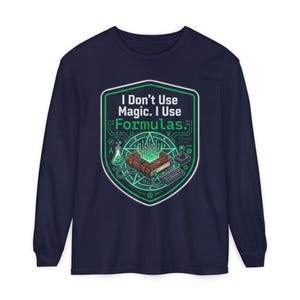 May include: A navy blue long-sleeve shirt featuring a graphic of a brown book with glowing green circuit lines rising from it. The design includes a flask, a keyboard, and a joystick, with the text "I Don't Use Magic. I Use Formulas."