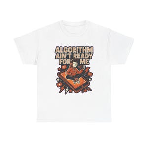 May include: A white t-shirt featuring a graphic of a person wearing headphones and holding a camera while sitting on a smartphone. The text ALGORITHM AIN'T READY FOR ME is printed above the illustration, which includes orange and black accents.