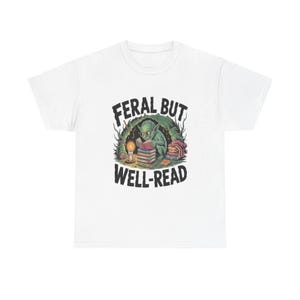 May include: A white t-shirt featuring a graphic illustration of a green goblin wearing glasses and reading a book while sitting on a stack of books. A lit candle sits nearby. The text FERAL BUT WELL-READ is printed in black above and below the goblin.