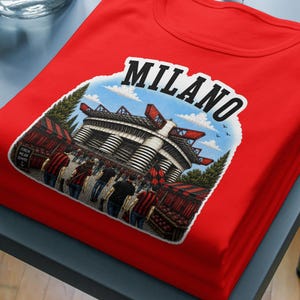Könnte beinhalten: Ein leuchtend rotes T-Shirt mit einem grafischen Aufdruck eines großen Stadions und dem darüber gewölbten Schriftzug MILANO. Die Illustration zeigt die Stadionfassade mit Bäumen und Personen im Vordergrund in einem detaillierten, farbigen Stil.