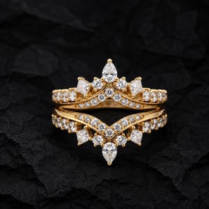 Könnte beinhalten: Ein Goldring-Set bestehend aus zwei ineinandergreifenden Bändern mit Chevron-Design. Die Bänder sind mit zahlreichen kleinen runden weißen Steinen besetzt, akzentuiert durch größere birnenförmige und quadratische weiße Steine.