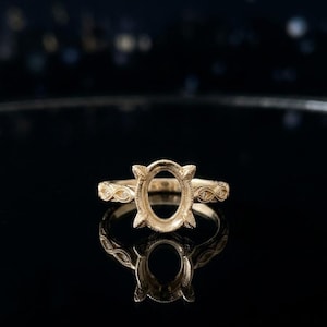 Könnte beinhalten: Ein goldener Ringrohling mit einer ovalen Fassung und vier Krappen. Die Ringschiene zeigt ein texturiertes, sich wiederholendes Unendlichkeitsmuster an den Seiten. Das Metall weist eine matte, körnige Oberfläche auf.