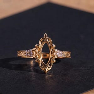 May include: A gold ring featuring an empty marquise-shaped setting in the center, surrounded by ornate scrollwork and small clear stones embedded along the band. The metal surface displays intricate engraved patterns on both sides of the setting.