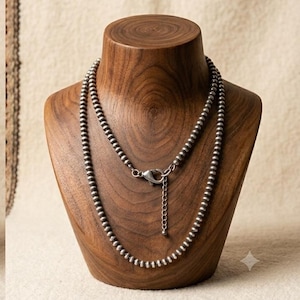 May include: A silver-toned beaded necklace featuring small, uniform spherical beads strung together. The necklace includes a metal lobster claw clasp and a short extender chain with a small bead at the end, displayed on a wooden neck form.