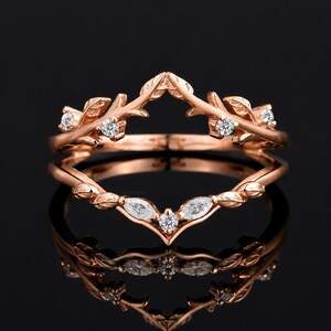 Può includere: Un coprianello in oro rosa con un design a forma di V, decorato con motivi a foglia e piccole pietre trasparenti a taglio rotondo e marquise. La fascia ha una finitura lucida ed è strutturata per incorniciare un altro anello con eleganza.