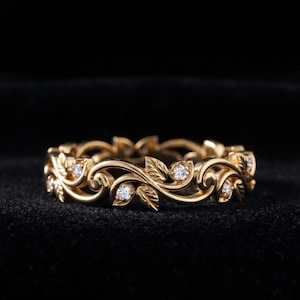 May include: A gold band ring featuring an intricate design of sculpted leaves and swirling vines. The metal surface is polished and yellow in tone. Several small, round, clear, faceted stones are set within the vine and leaf pattern.