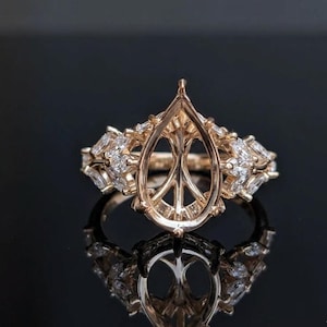 May include: A rose gold ring setting featuring a central pear-shaped prong mount designed to hold a gemstone. The band is adorned with multiple marquise-cut clear stones arranged in a cluster pattern on both sides of the central setting.