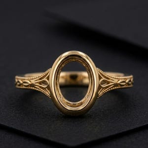 May include: A polished yellow gold ring setting featuring a central oval-shaped bezel designed to hold a gemstone. The band displays intricate, symmetrical filigree metalwork along the shoulders, extending from the central oval frame.