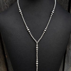 Puede incluir: Un collar en forma de Y que presenta una serie de cuentas plateadas pulidas. Las cuentas varían en tamaño, con esferas más grandes situadas cerca de la parte superior y cuentas más pequeñas que descienden hacia el centro, creando un diseño metálico graduado.