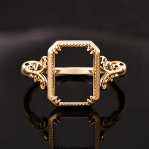 May include: A gold ring featuring a rectangular setting with four prongs at the corners. The band is decorated with intricate scrollwork patterns on both sides of the central frame, which has a beaded texture along its outer edges.