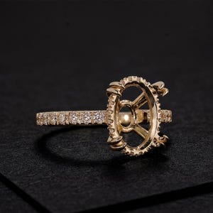 May include: A gold ring setting featuring an oval-shaped center mount with four prongs. The band and the outer edge of the center mount are encrusted with small, round, clear pavé-set stones that catch the light, creating a sparkling appearance.