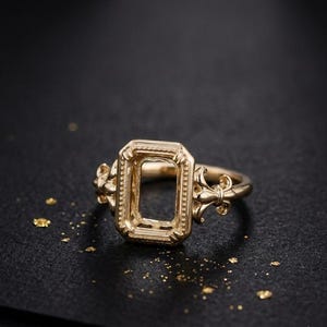 Könnte beinhalten: Ein goldfarbener Ring mit einer rechteckigen, leeren Fassung und einem perlenartigen Rand. Die Seiten des Rings sind mit dekorativen Fleur-de-Lis-Motiven verziert. Die Ringschiene ist glatt und rund und für einen Edelstein in der Mitte ausgelegt.