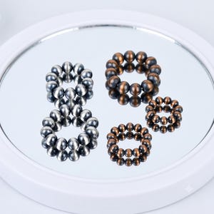 Könnte beinhalten: Vier kreisförmige Ringe aus kleinen, kugelförmigen Perlen. Zwei Ringe haben ein poliertes silberfarbenes Finish, während die anderen beiden Ringe ein dunkles, mattes bronze- oder kupferfarbenes Finish aufweisen. Jeder Ring besteht aus einer Perlenkette.