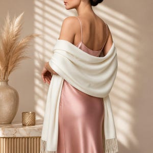 May include: A smooth, solid cream-colored shawl with fringe trim draped over a satin slip dress. The shawl is worn over the shoulders, covering the back and upper arms, while the dress features thin spaghetti straps and a soft pink sheen.