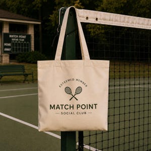 Puede incluir: Bolso tote de lona beige con el texto "ESTEEMED MEMBER" sobre raquetas de tenis cruzadas y "MATCH POINT SOCIAL CLUB" debajo. El bolso cuelga de un poste de red de tenis en una cancha de tenis.