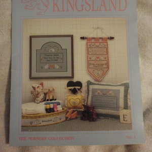 May include: A nursery themed cross stitch pattern booklet with a blue cover and the title "KINGSLAND" in pink lettering. The cover features a cross stitch design of a lion with a crown. The booklet includes a variety of cross stitch patterns for baby items, including a baby blanket, a baby quilt, and a baby bib. The booklet also includes instructions for how to stitch the patterns.