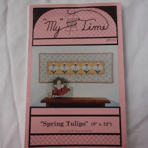 May include: A craft pattern in pink and white, featuring the text "My Time" and "Spring Tulips." The pattern includes a tulip design and a doll. The dimensions are 23 cm x 56 cm.
