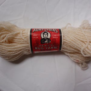 May include: A skein of off-white heavy rug yarn with a red and black paper label. The label features the text Aunt Lydia's, Fast Color, 70 Yards, and Heavy Rug Yarn, along with an illustration of a person in a dark collared shirt.