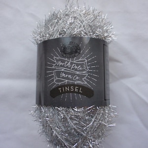 May include: A ball of silver tinsel yarn with a dark gray label. The label reads "North Pole Yarn Co." and "Tinsel" in white lettering. The yarn is fluffy and metallic, perfect for holiday crafts and decorations.